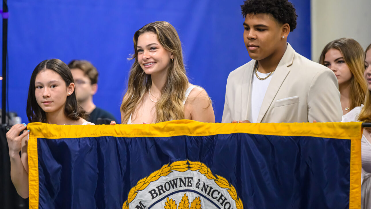 Three students, two females and one male, hold a banner with a school emblem in front of a blue background during what appears to be a formal event.