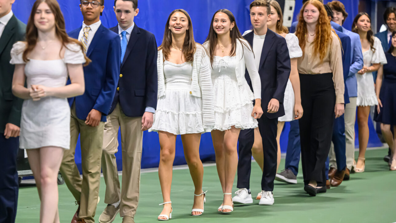 A group of young people dressed in semi-formal attire walk in a line indoors. Some are smiling and conversing.