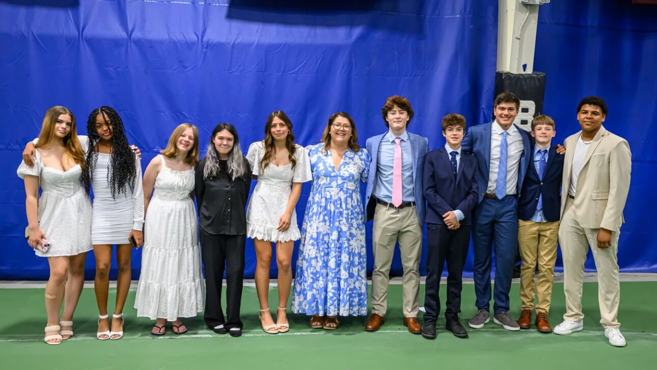 A group of eleven people, dressed in formal attire, stands in a line indoors against a blue background, smiling at the camera.