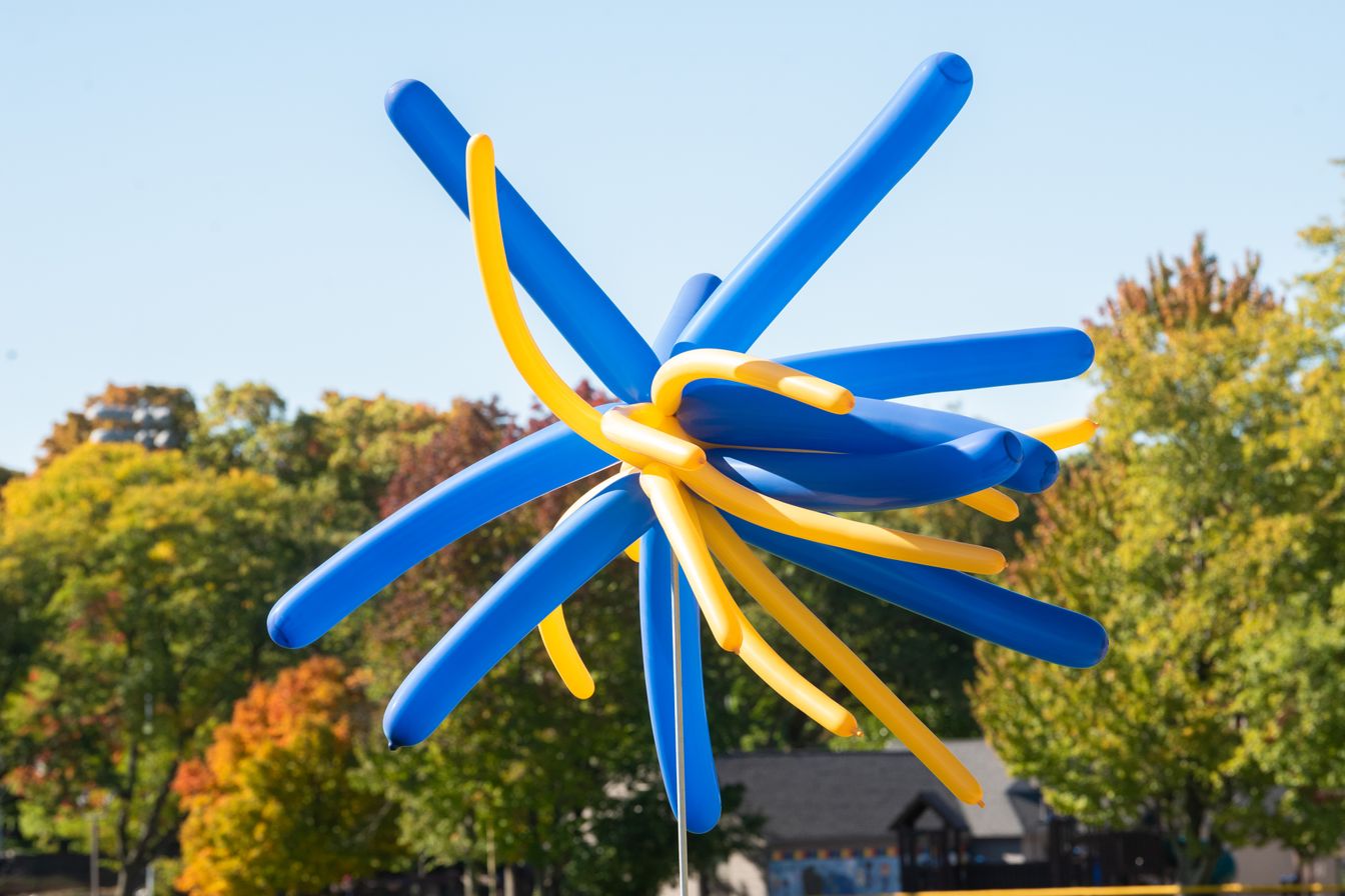 A cluster of blue and yellow balloons twisted together, resembling a starburst shape, set outdoors with trees in the background.